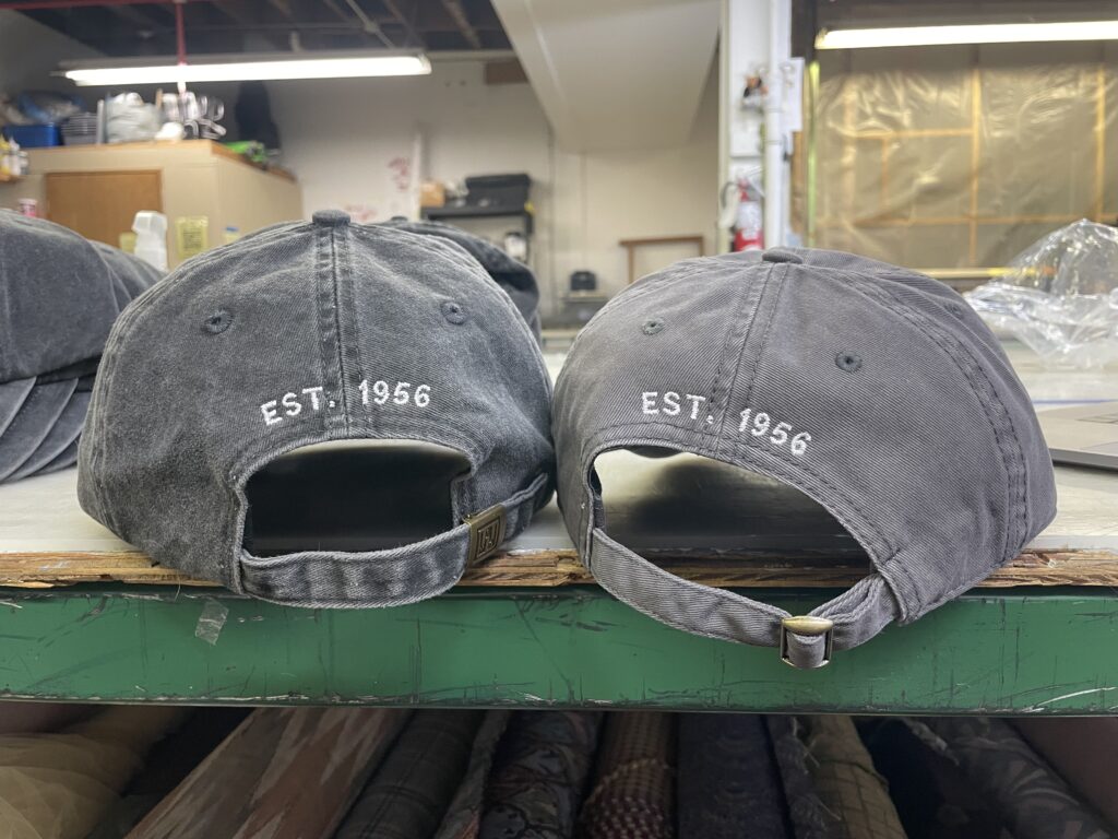 The back of two matching gray dad hats with EST. 1956 custom embroidery over the back in white thread
